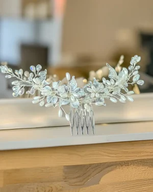 Tocado de novia en resina Coral con perlas y cristales Tocado de novia en resina con diseño floral que combina perlas y cristales para lograr un acabado delicado y luminoso. El modelo Coral se caracteriza por sus flores y pequeñas hojas que aportan un efecto sutil y elegante, ideal para realzar el peinado sin sobrecargarlo. Su brillo suave crea un equilibrio perfecto entre sofisticación y naturalidad, convirtiéndolo en un accesorio ideal para novias que buscan un look refinado. Su estructura flexible permite adaptarlo fácilmente a distintos estilos de peinado: Recogidos elegantes Semirecogidos románticos Peinados sueltos con ondas Puede ubicarse en diferentes zonas del cabello, logrando un acabado armónico que acompaña el estilo sin competir con otros elementos del look. Su diseño liviano facilita su uso durante toda la celebración, brindando comodidad y firmeza. Cada pieza presenta ligeras variaciones en sus detalles, lo que aporta un carácter único a cada accesorio. 📏 Detalles Material: Resina, perlas y cristales Estilo: Floral / Elegante / Romántico Uso: Novia Medidas: 22 cm aprox. Puede existir una pequeña variación en el tamaño.